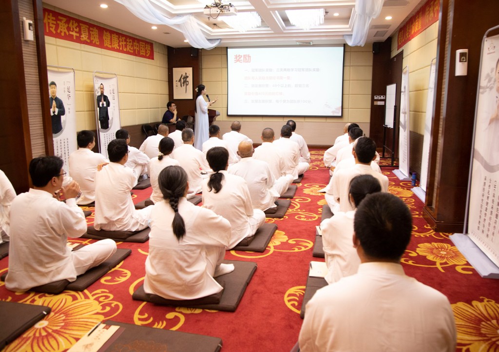 A group of people in white attire seated on mats in a conference room, attentively listening to a presentation displayed on a screen.