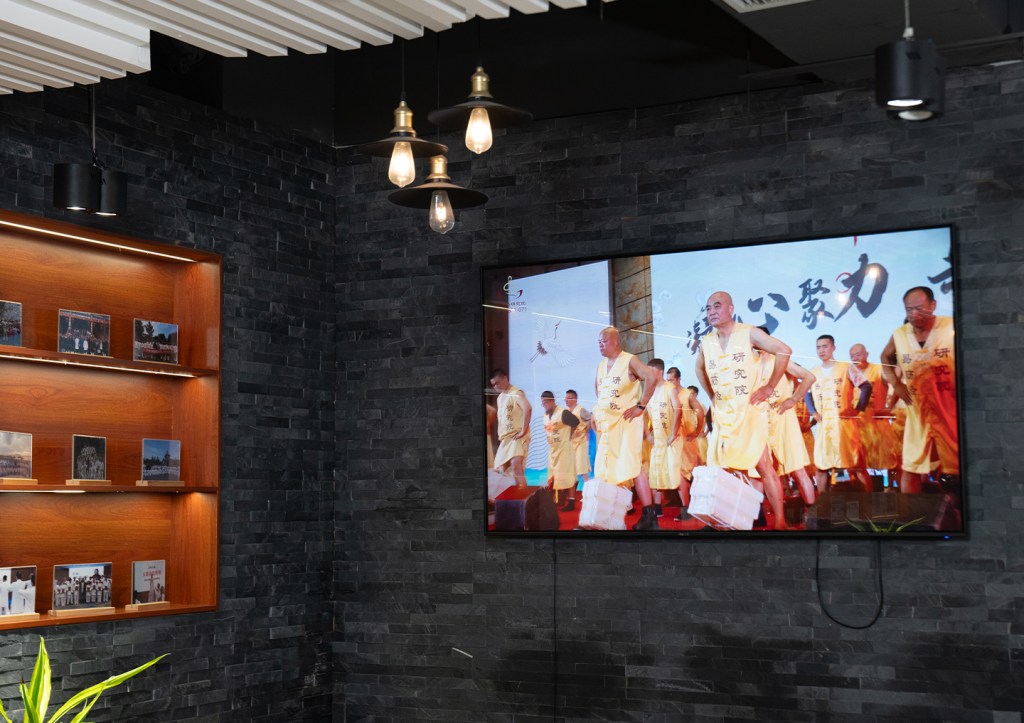 A modern interior space featuring a television displaying a performance by individuals in yellow costumes, surrounded by black stone walls and a wooden display shelf with framed photographs.