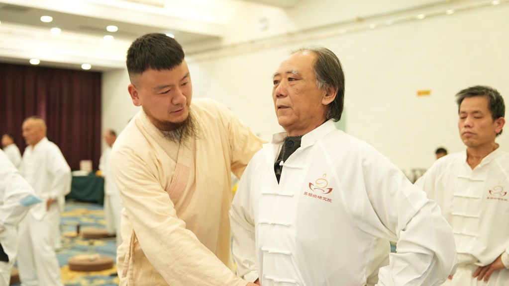 A practitioner is demonstrating a technique on an elderly man during a training session, with other participants practicing in the background.