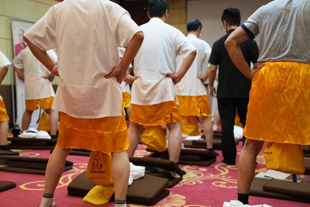 A group of individuals in white shirts and orange shorts, standing in a fitness or training setting, with their hands on their hips.