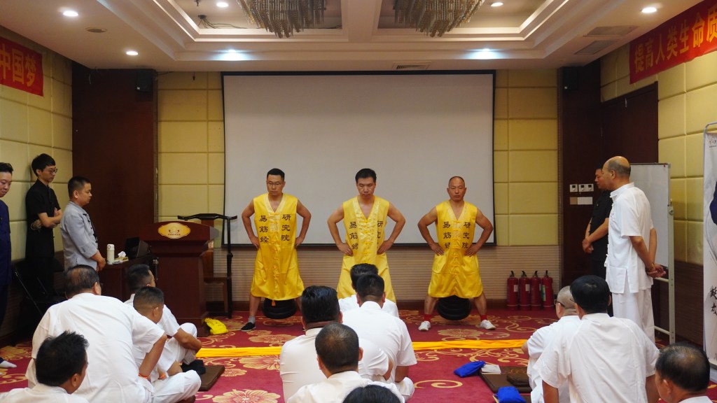 Three performers in yellow attire demonstrate martial arts poses in front of an audience in a conference room.