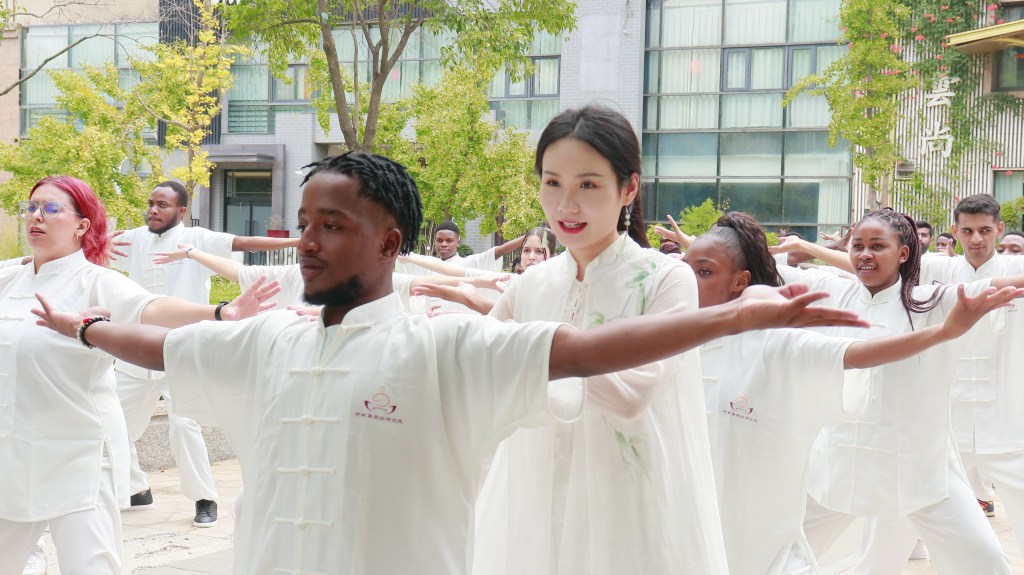 Group of individuals practicing Tai Chi in a park setting, wearing traditional white attire. The scene features a mix of participants, including a woman leading the group with graceful movements.